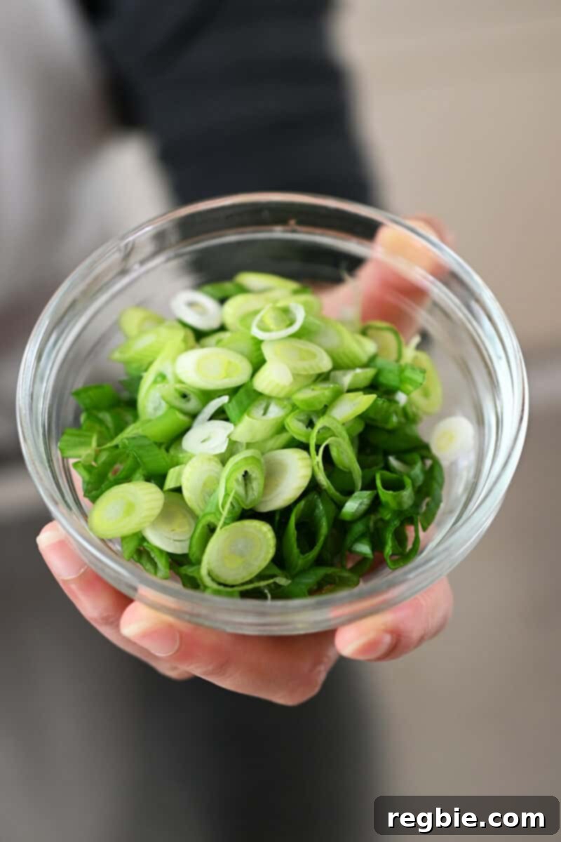 A hand is presenting a small glass bowl filled with finely thinly sliced scallions, prepared for garnish.