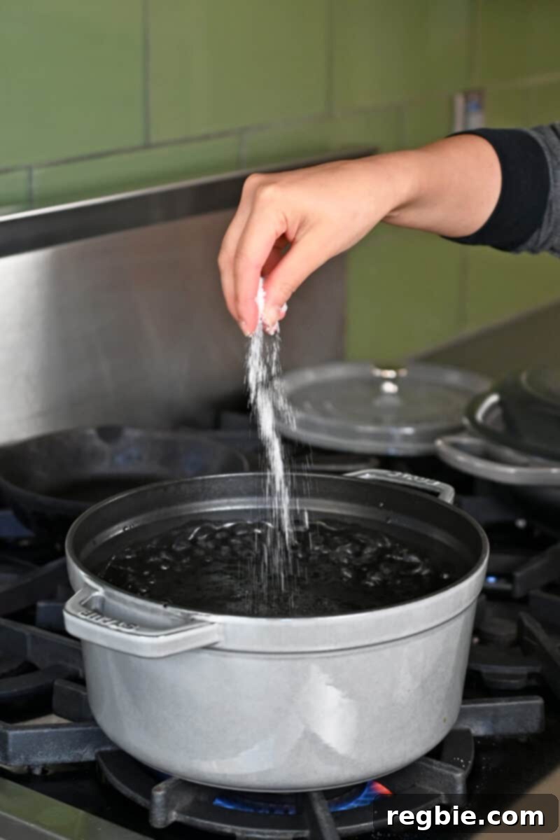 A hand is adding a pinch of salt into a large stockpot filled with boiling water, preparing to blanch vegetables.