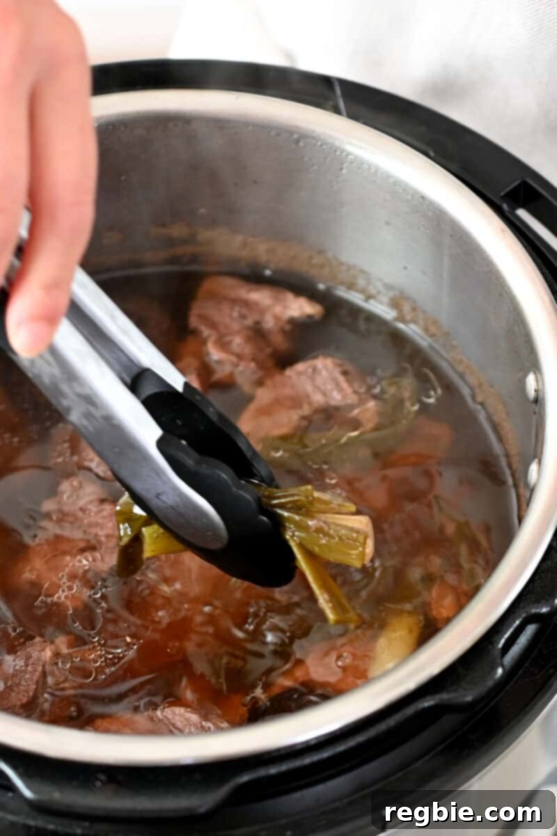 A pair of tongs is removing sliced ginger from an open Instant Pot filled with richly flavored beef broth.