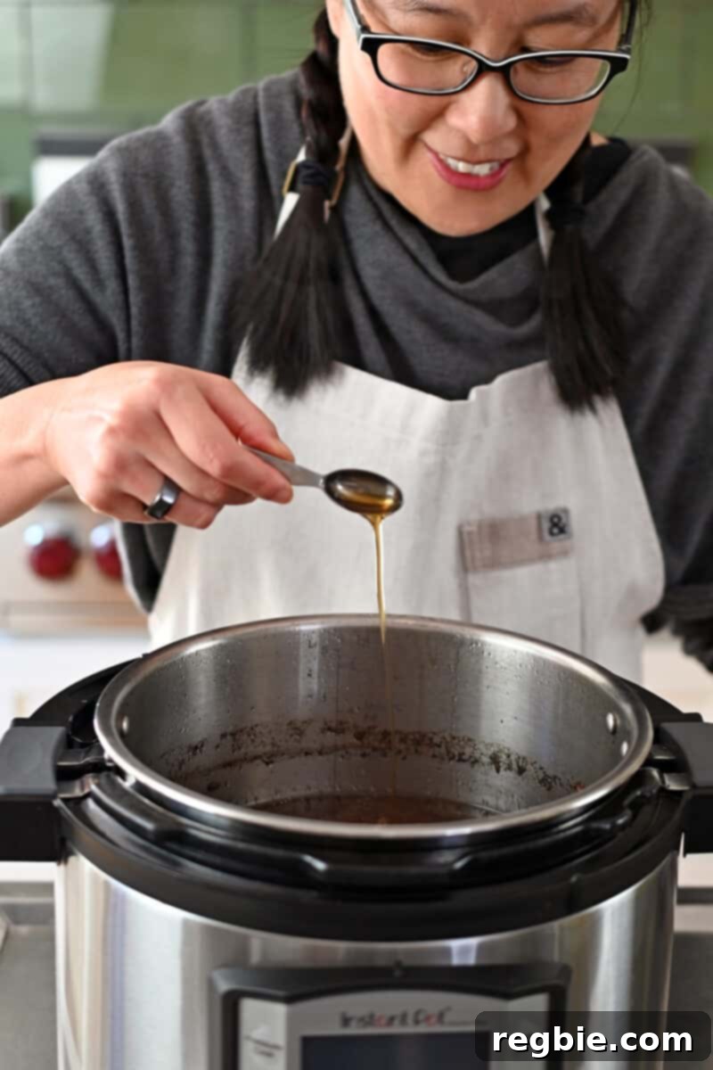 A smiling Asian woman is adding a spoonful of toasted sesame oil to an Instant Pot filled with delicious beef broth.