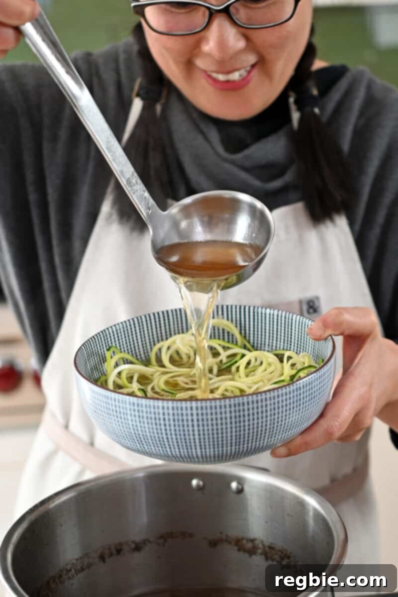 A smiling Asian woman is ladling hot beef broth onto a bowl of spiralized zucchini noodles, preparing a delicious soup.