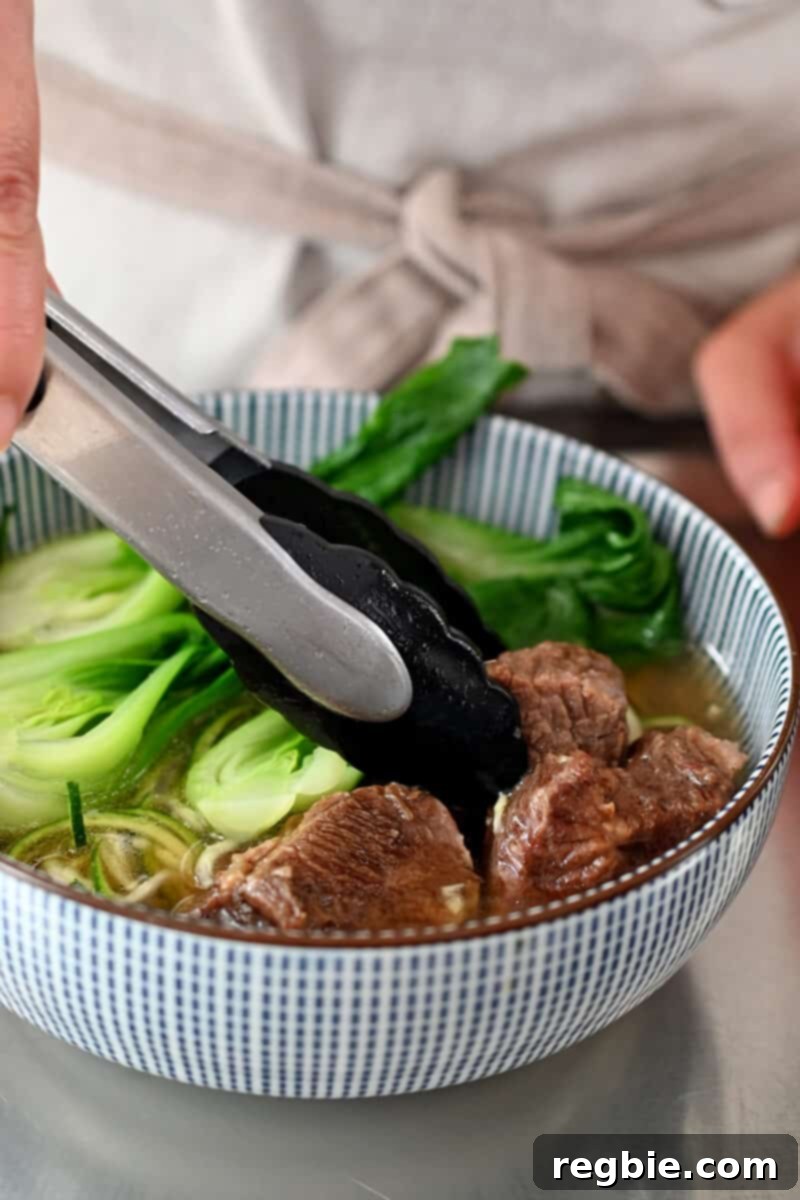 A pair of tongs is adding tender stewed beef cubes on top of a bowl of Chinese beef noodle soup, ready for garnish.