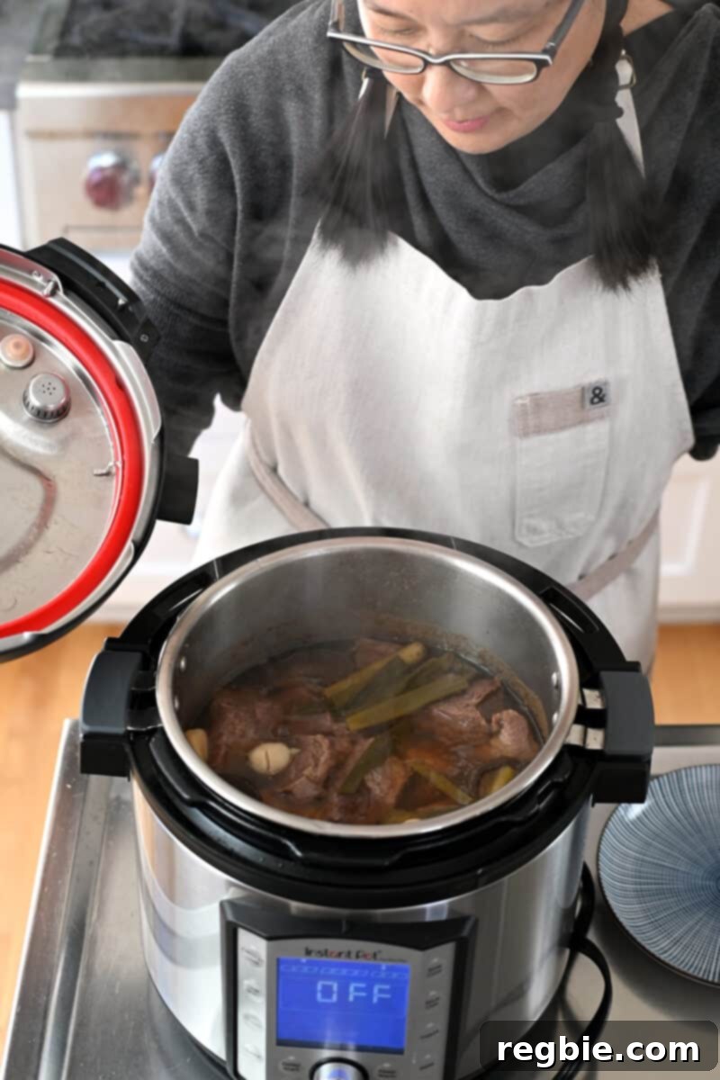 An Asian woman taking the lid off an Instant Pot filled with steaming Chinese Beef Noodle soup, steam rising.