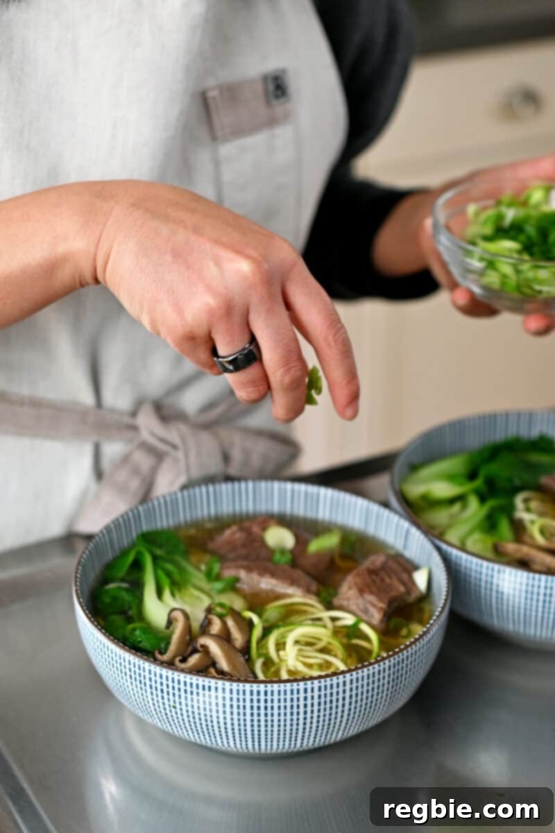 A hand is sprinkling finely sliced scallions over a bowl of paleo, Whole30, gluten-free, and keto Chinese Beef Noodle Soup, ready to serve.