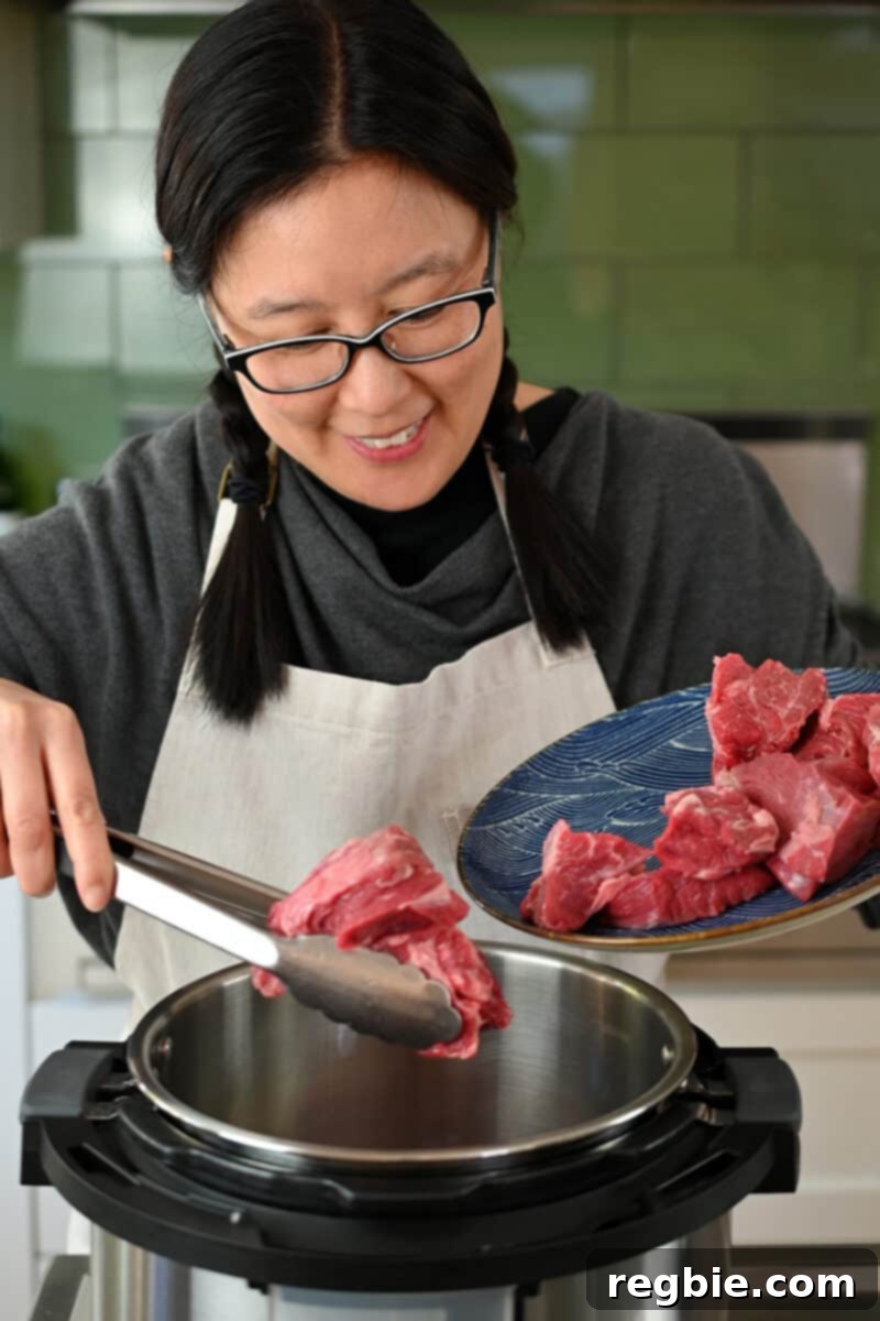 An Asian woman in glasses is adding beef cubes to an open Instant Pot, preparing to make Chinese Beef Noodle Soup.