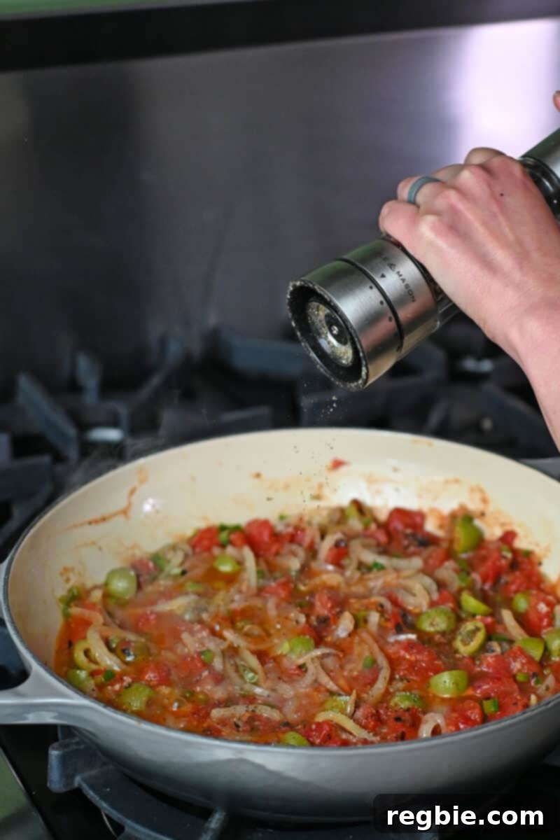 A hand grinds fresh black pepper over a skillet of homemade Veracruz sauce, adding a final touch of seasoning to perfect its taste.