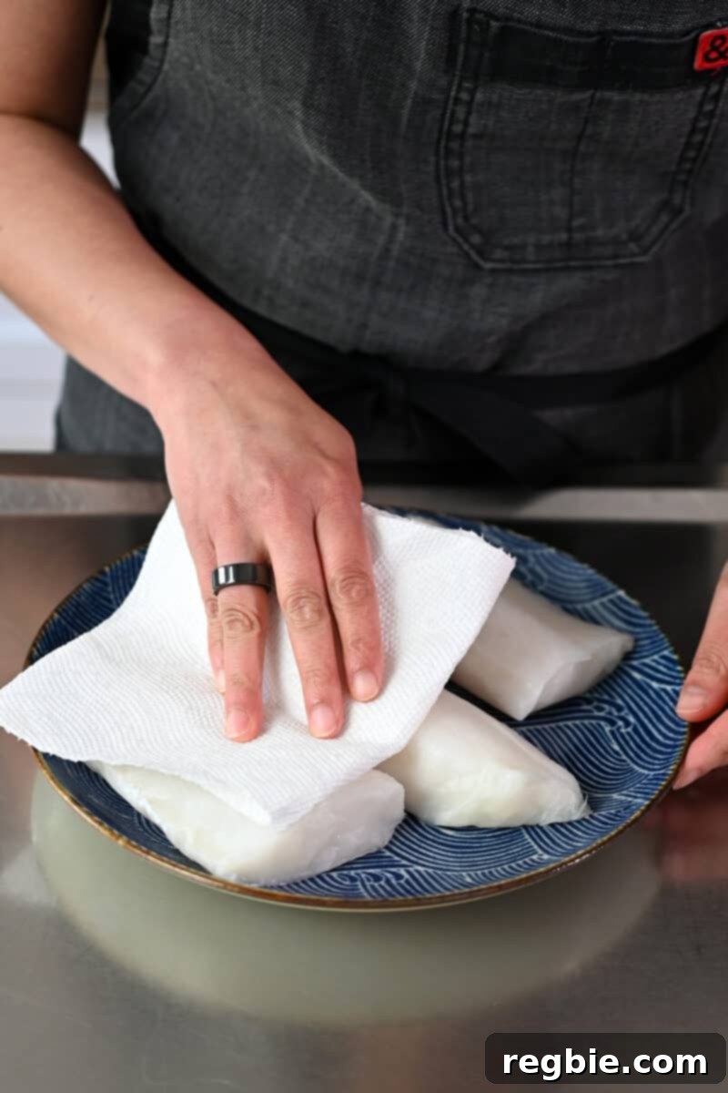 A chef's hand gently pats raw white fish fillets dry with a paper towel, a crucial step before seasoning them thoroughly with salt and pepper.