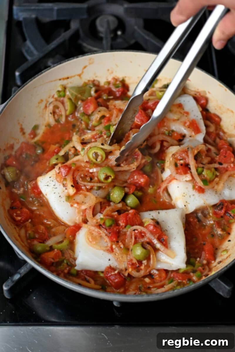 A pair of tongs carefully places raw fish fillets into a skillet brimming with rich Veracruz sauce, preparing them for a gentle simmer.
