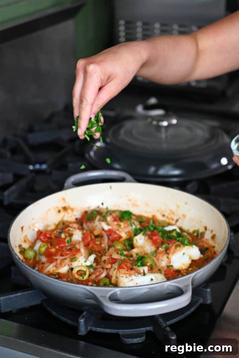 A hand elegantly sprinkles vibrant green minced fresh cilantro over a pan of perfectly cooked white fish in Veracruz sauce, adding a final touch of freshness.
