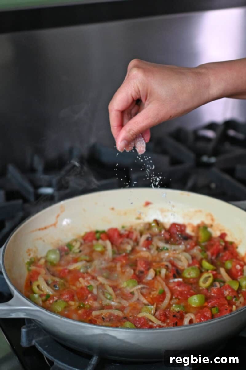 A hand sprinkles kosher salt into a simmering skillet of bright red Veracruz sauce, enhancing its deep flavors before the fish is added.