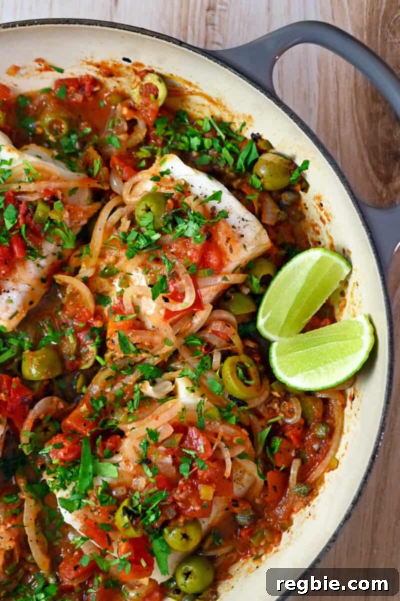 An inviting overhead shot of perfectly cooked white fish in Veracruz sauce nestled in a skillet, accompanied by bright, refreshing sliced lime wedges.