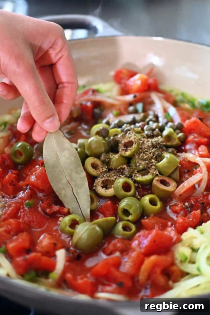 A hand carefully adds a single dried bay leaf to a vibrant mixture of fire-roasted diced tomatoes, green olives, capers, and dried oregano simmering in a skillet, creating the authentic Veracruz sauce.