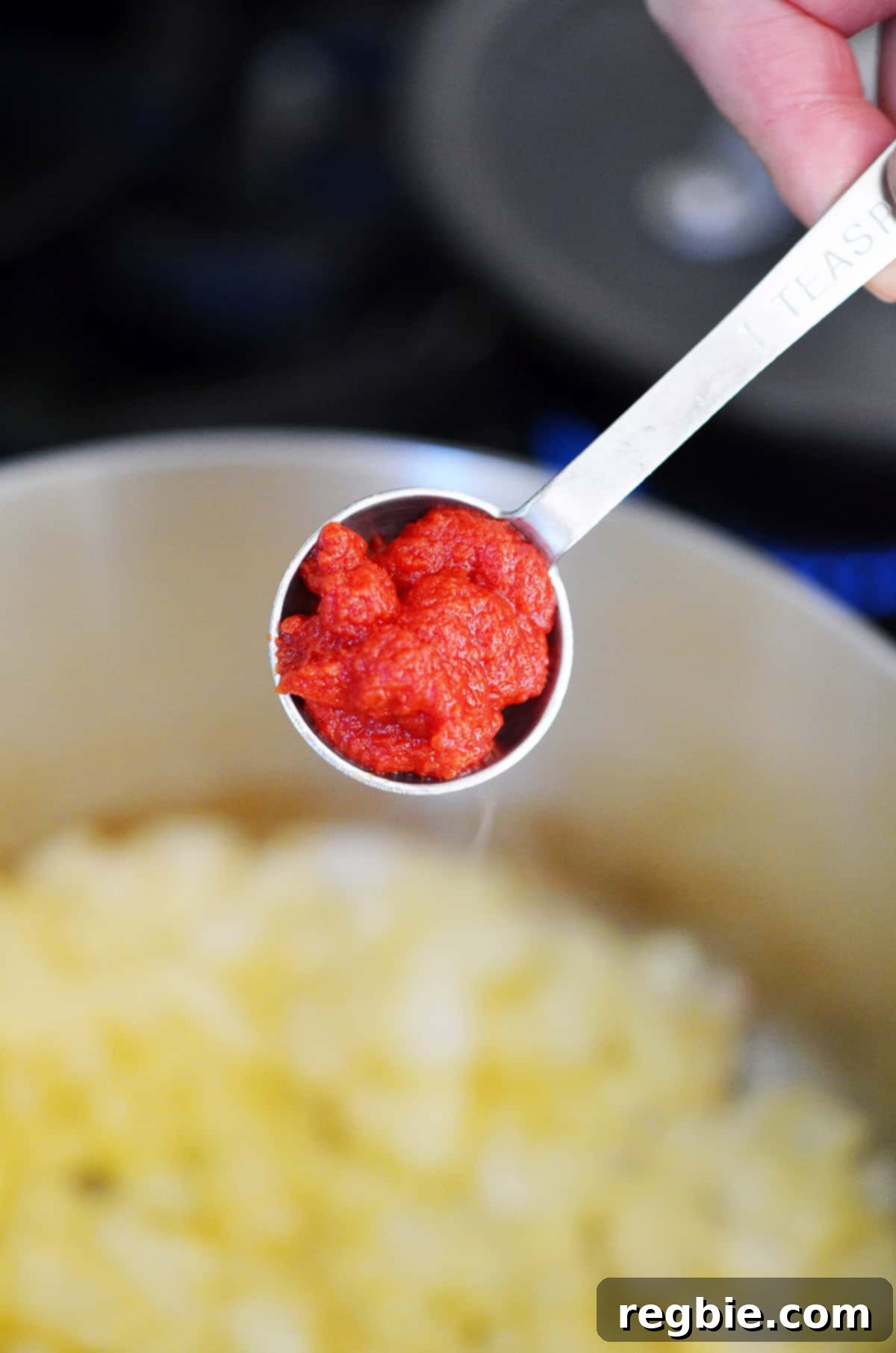 Adding a spoonful of tomato paste to a saucepan filled with sautéed diced onions.
