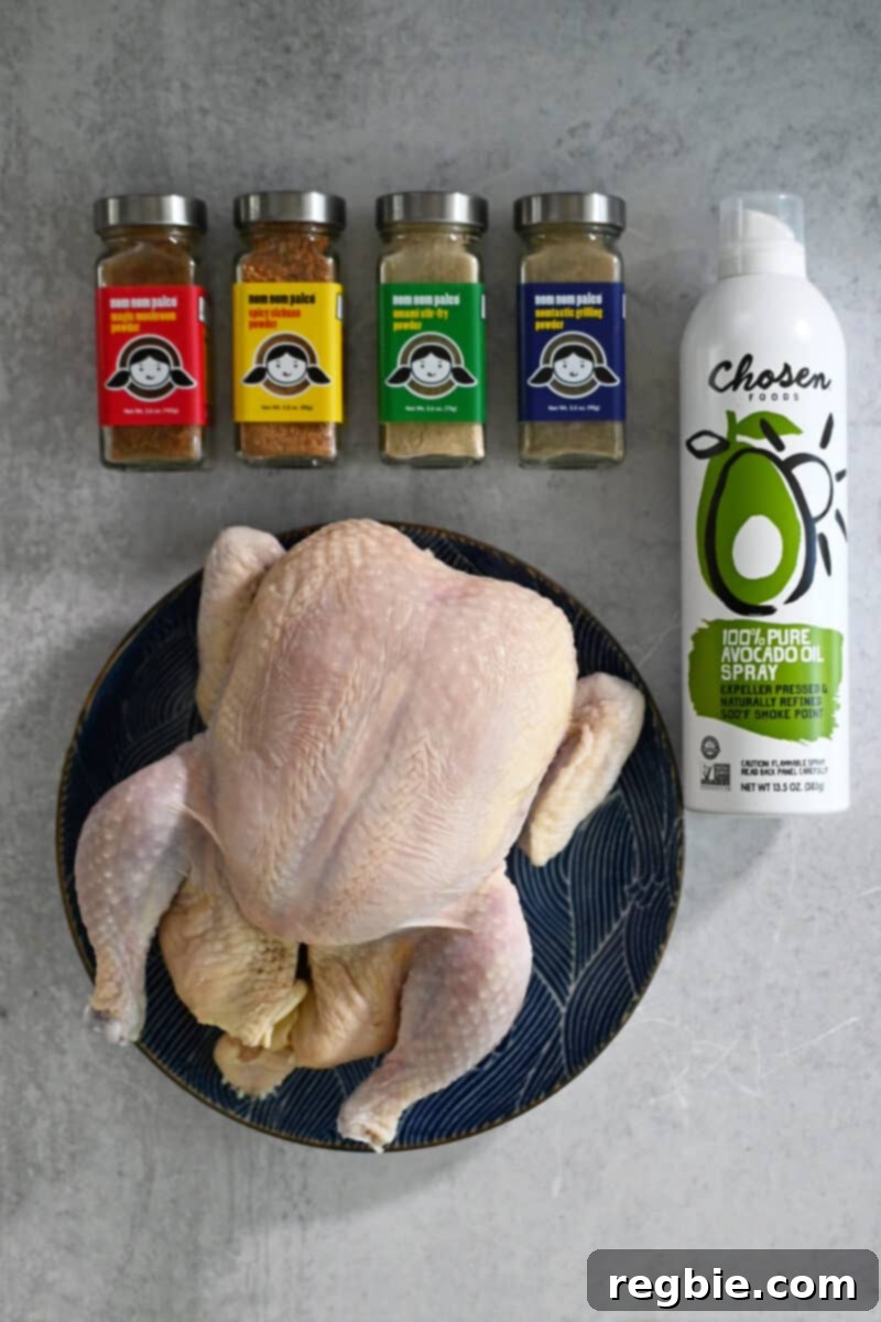 Golden Crispy Air Fried Whole Chicken 4 An overhead shot of the raw ingredients for air fryer whole chicken: Nom Nom Paleo spice blends, a whole raw chicken, and avocado oil spray, laid out on a clean surface.