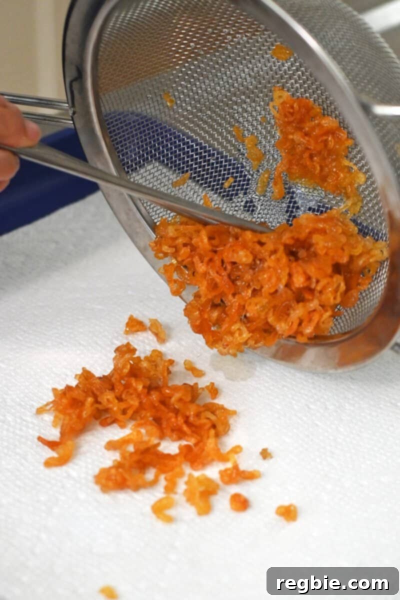 Crispy shallots are being transferred from a sieve to a paper towel-lined rimmed baking sheet for draining.