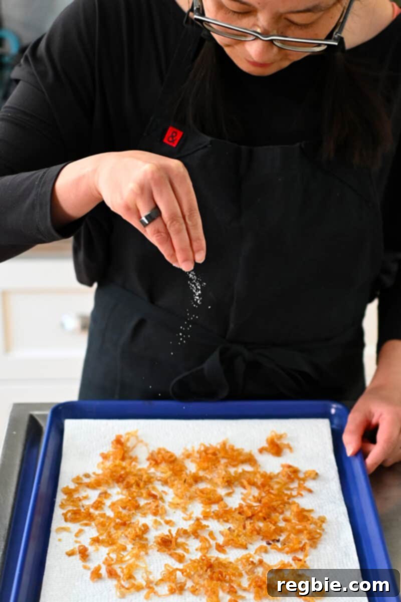 An Asian woman in a black apron is delicately sprinkling salt onto a fresh batch of hot crispy shallots spread on a tray.