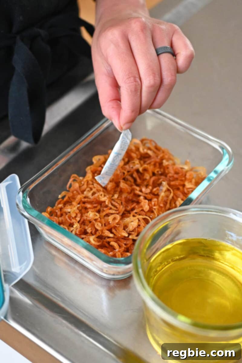 A hand is carefully adding a desiccant packet to a clear glass container filled with crispy shallots to help preserve their crunch.