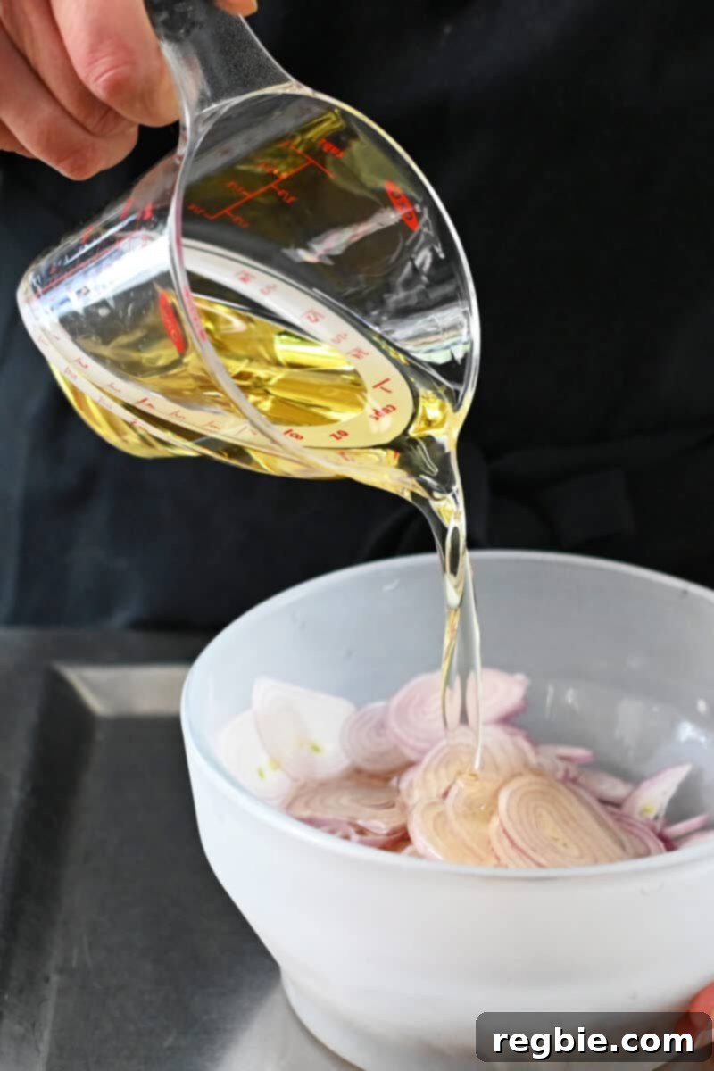 Avocado oil is being poured from a liquid measuring cup into a glass bowl containing sliced shallots, ensuring full coverage.