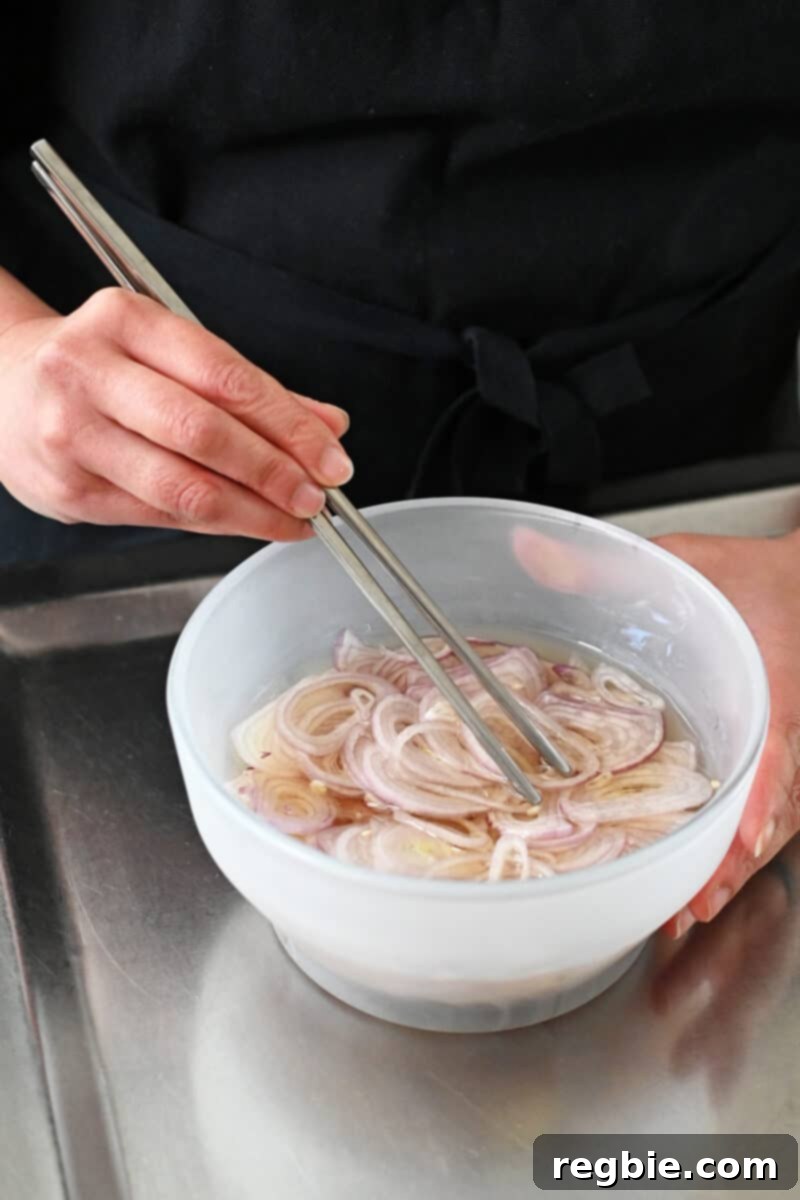 Someone is using stainless steel chopsticks to gently submerge sliced shallots into the oil within a glass bowl.