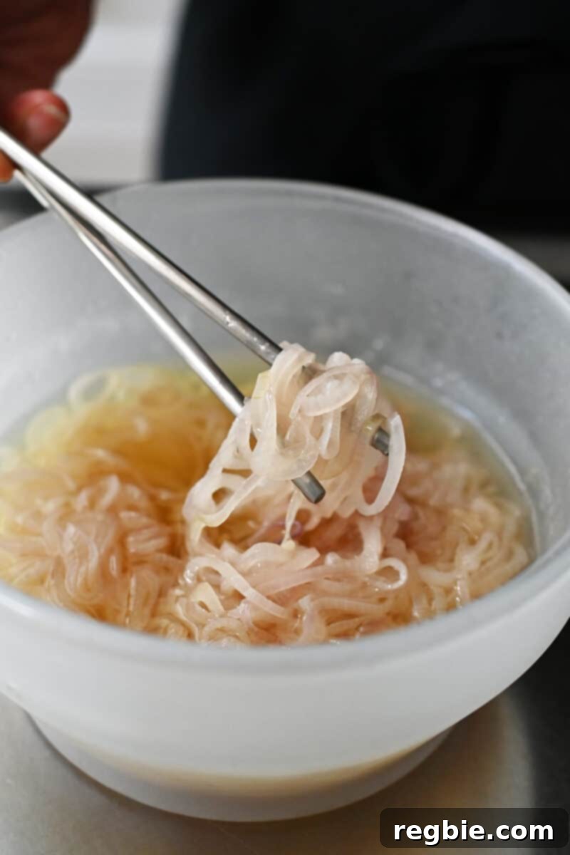 A pair of chopsticks delicately lifts softened sliced shallots from a bowl of avocado oil, showing their initial cooking stage.