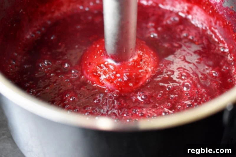 Someone using an immersion blender to mix the ingredients for paleo cran-cherry sauce together, illustrating how to achieve a smoother texture.