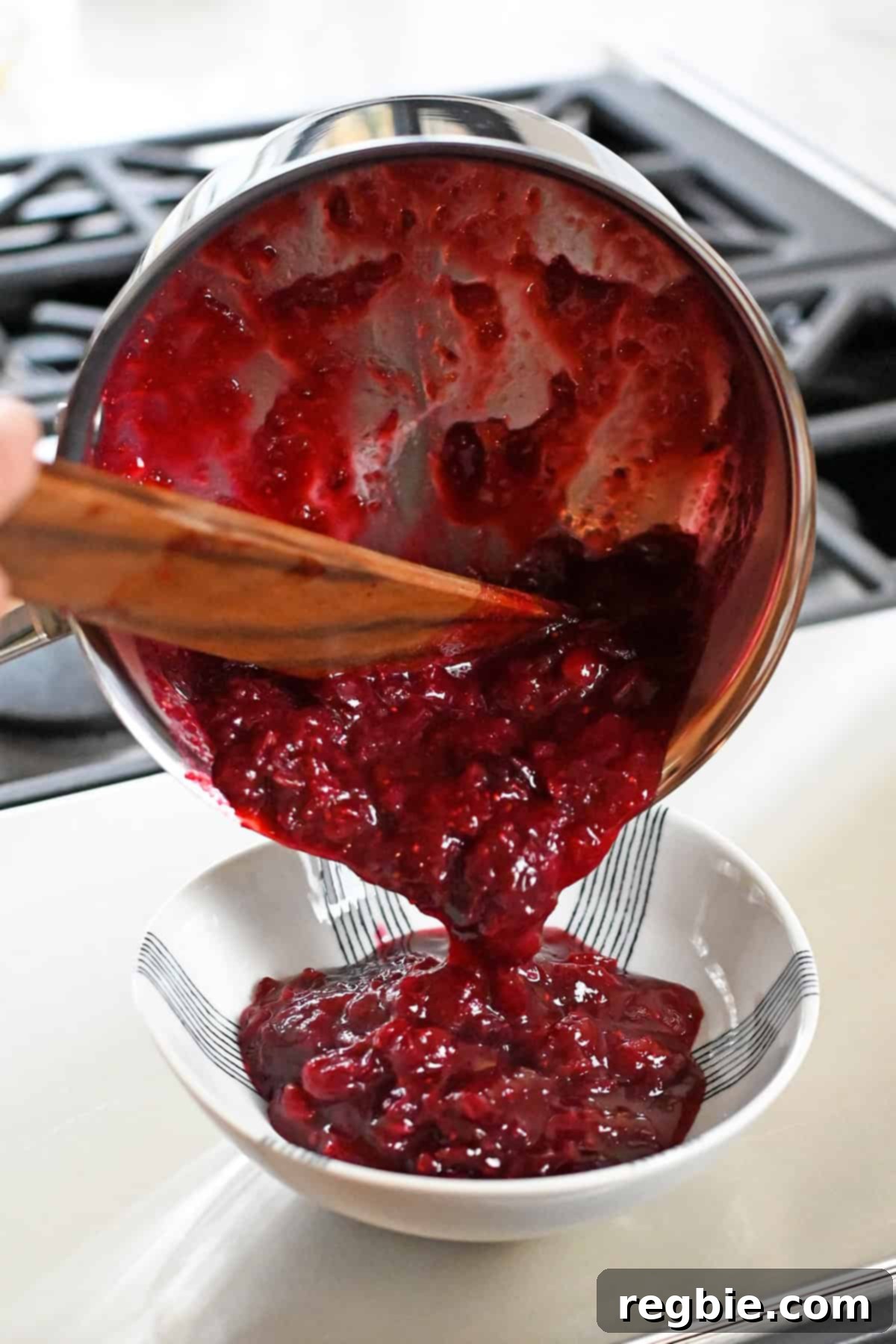 Pouring paleo cranberry sauce from a saucepan into a white serving bowl, ready to be presented.