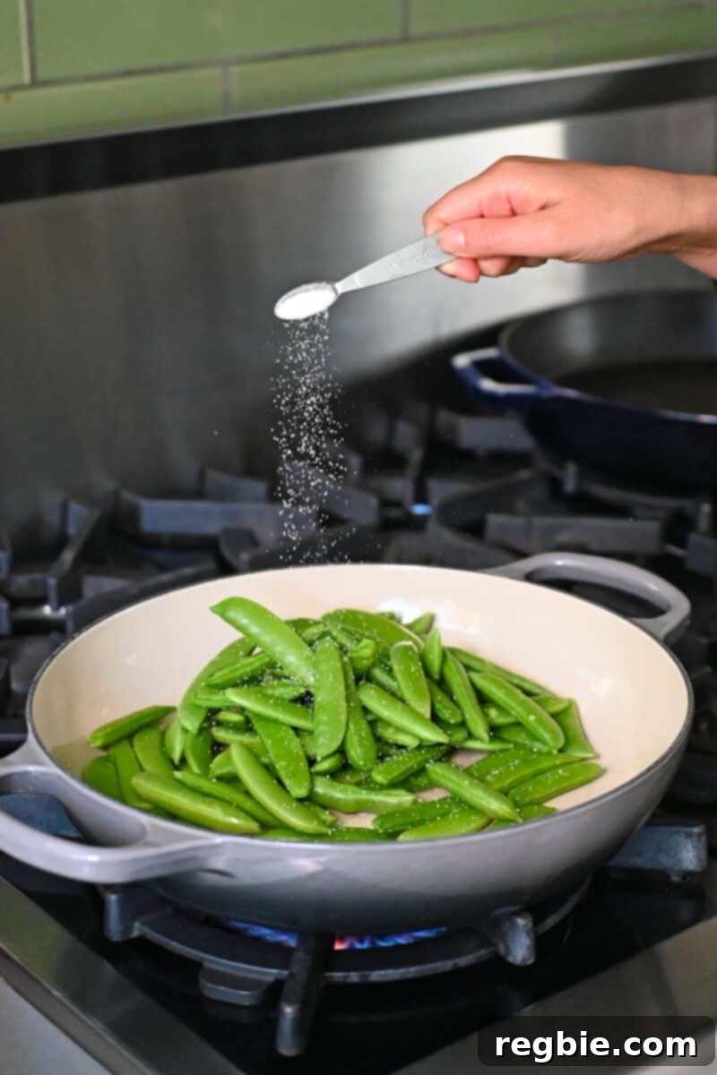 Speedy Southeast Asian Snap Pea Sensation 6 Adding a small spoonful of Diamond Crystal kosher salt into a skillet with vibrant green sugar snap peas.