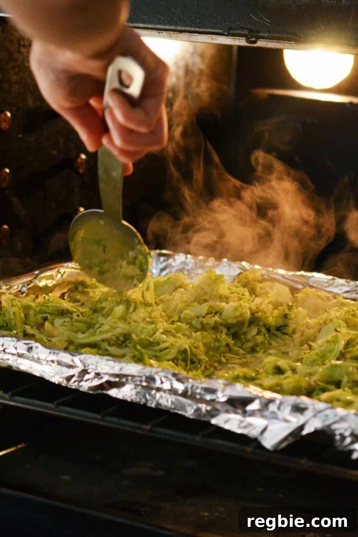 A large metal spoon stirring some roasted shaved Brussels sprouts on a foil-lined rimmed baking sheet inside the oven, showing the progress of browning.
