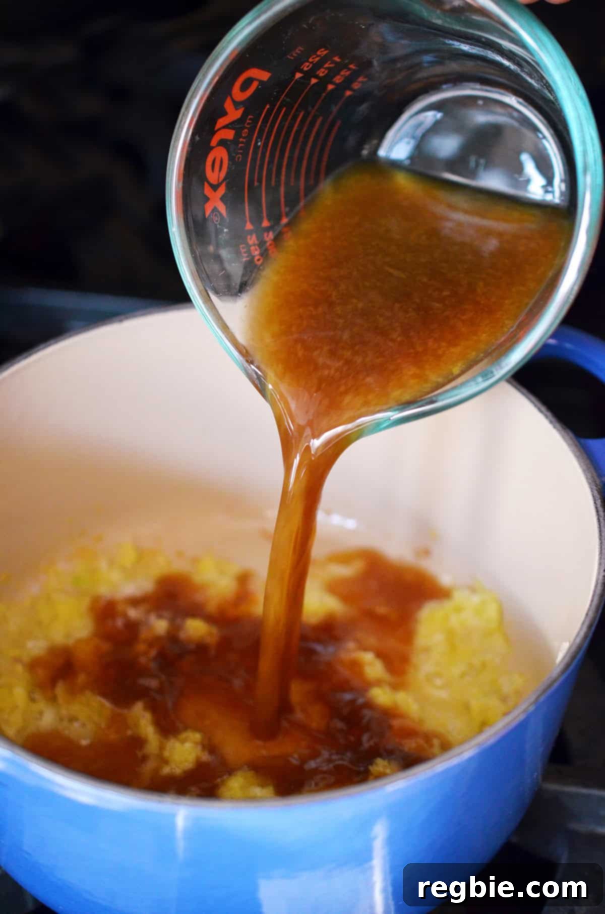 Pouring a brown liquid, representing the mix of orange juice, coconut aminos, rice vinegar, and fish sauce, into a blue saucepan containing sautéed ginger, garlic, and shallots.