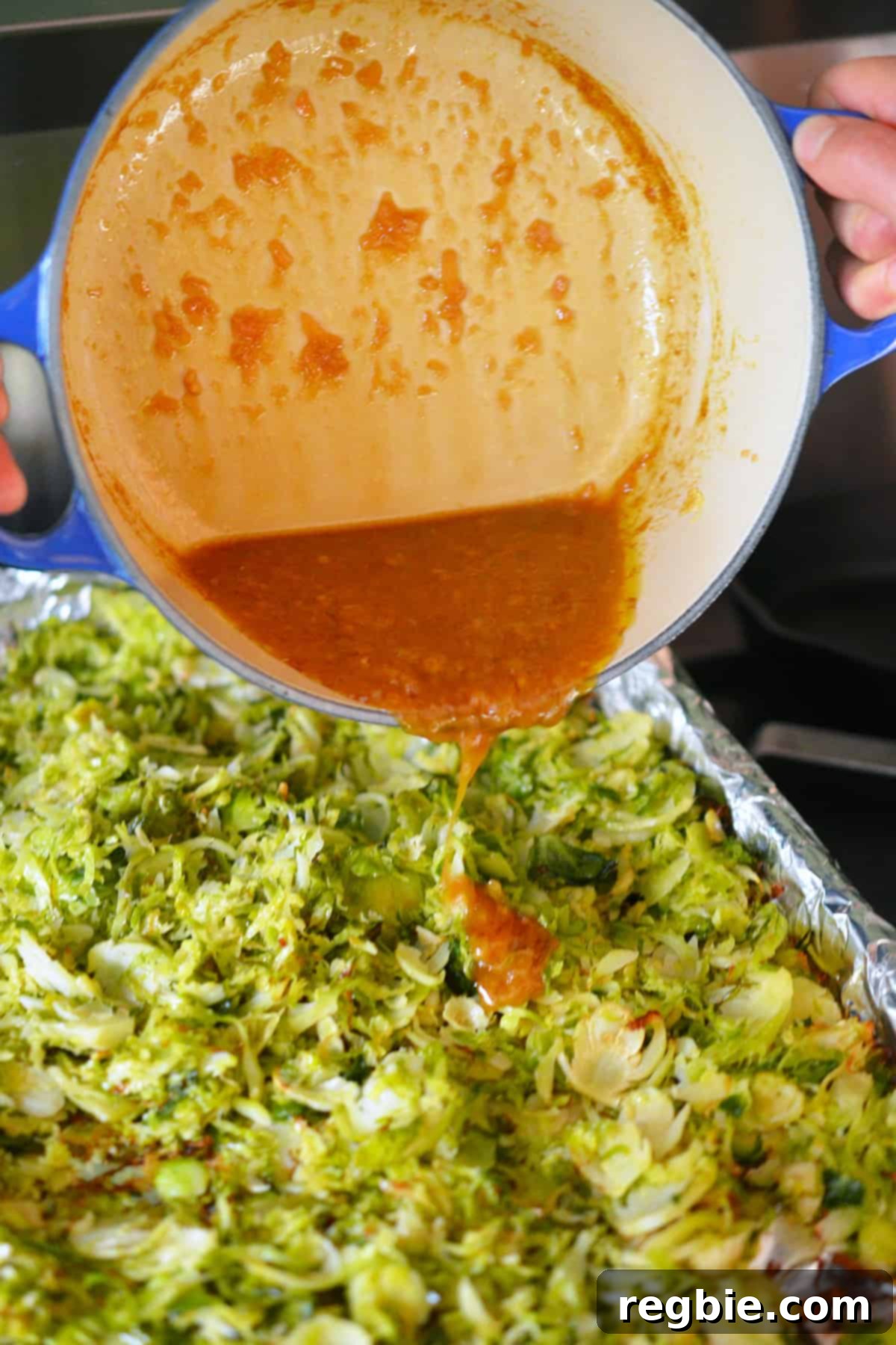 Pouring the vibrant Asian citrus dressing from a blue saucepan directly onto a rimmed baking sheet filled with freshly roasted shaved Brussels sprouts, infusing them with flavor.