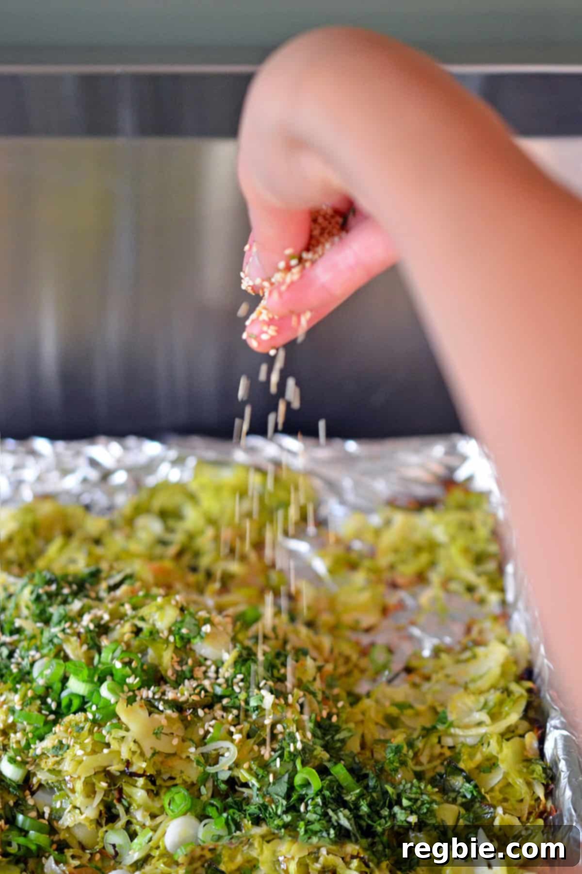 A hand sprinkling toasted sesame seeds over a rimmed baking sheet generously filled with roasted shaved Brussels sprouts and fresh herbs, adding the final touch before serving.