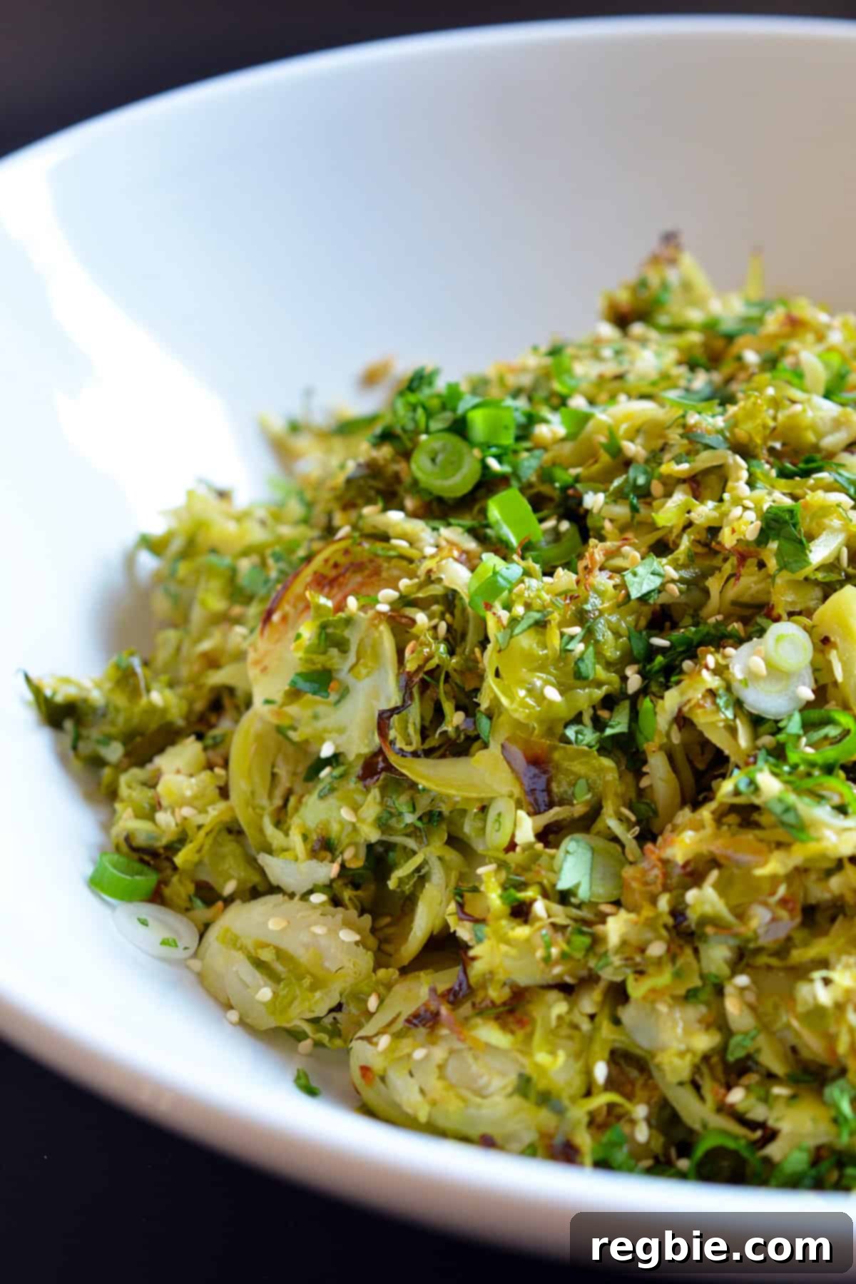A closeup of a large white serving bowl filled with a colorful Roasted Brussels Sprouts Salad with Asian Citrus Dressing, garnished with fresh herbs and sesame seeds.