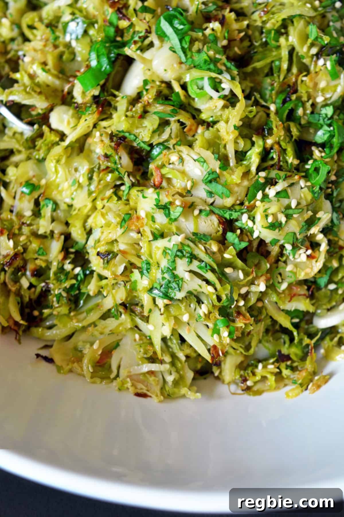 An overhead closeup shot of a white bowl filled with Roasted Brussels sprouts salad with Asian dressing, beautifully topped with thinly sliced green onions and toasted sesame seeds, ready to be served.