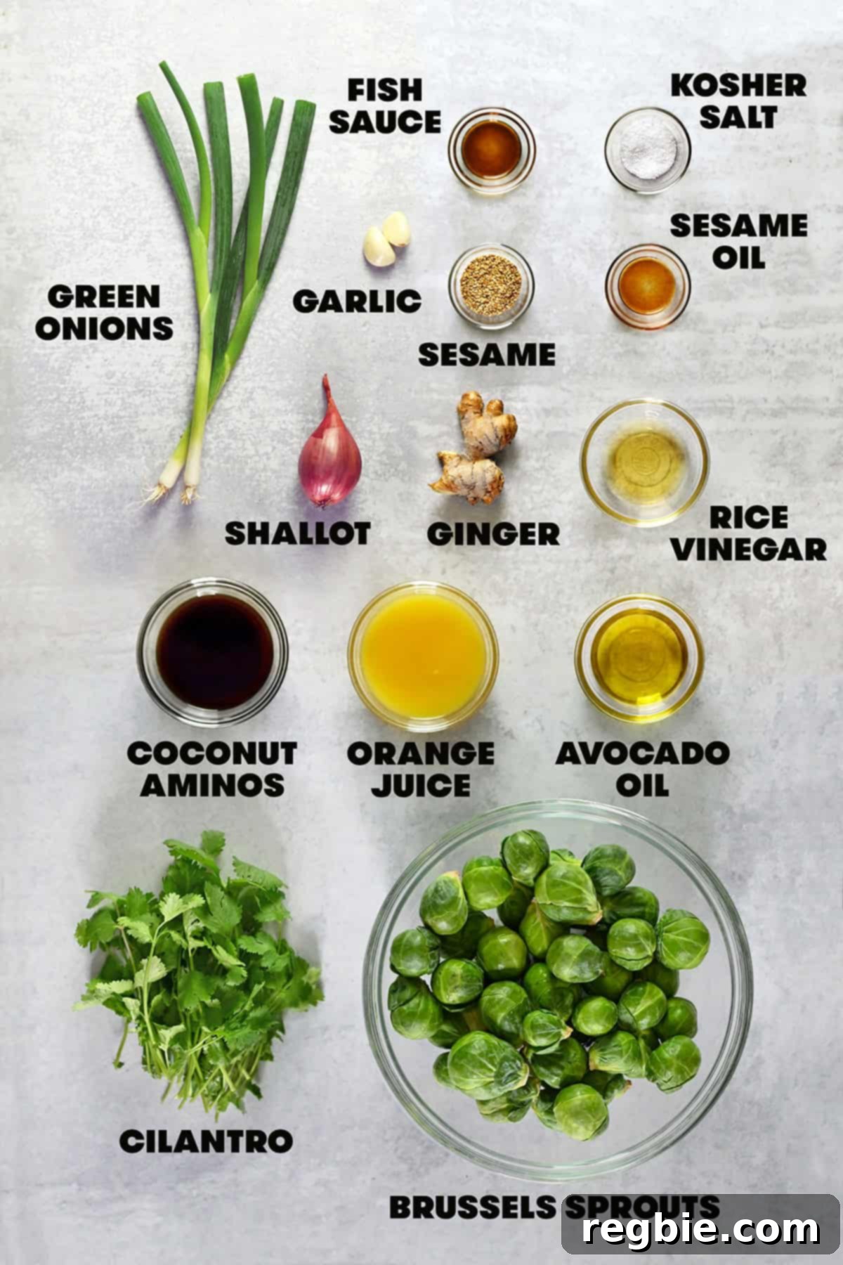 An overhead shot of the raw ingredients laid out on a clean surface, ready to make roasted Brussels sprouts salad with Asian citrus dressing, including fresh Brussels sprouts, ginger, garlic, shallots, and various liquids.