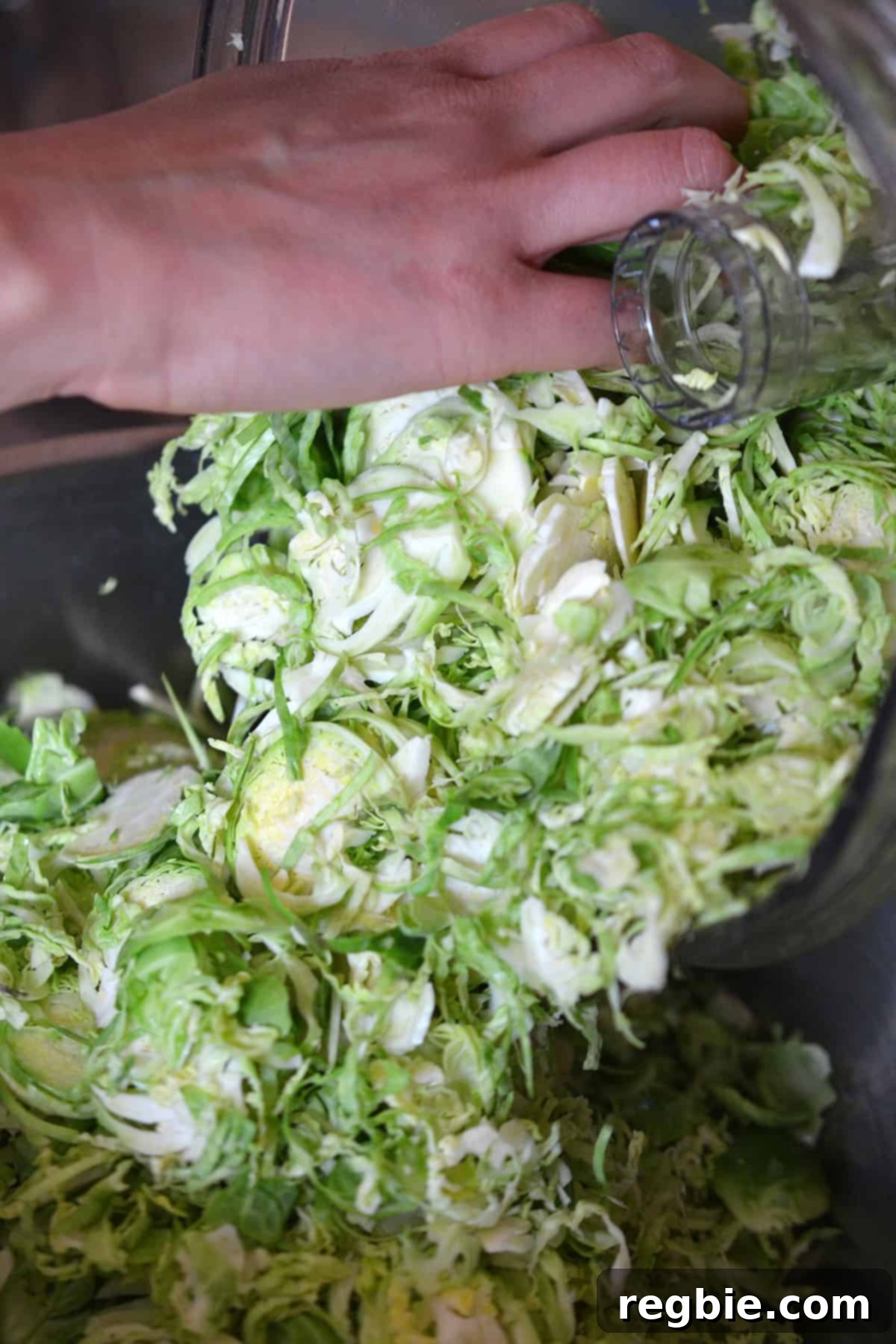 The process of pouring the freshly shaved Brussels sprouts out of a food processor and into a large mixing bowl, illustrating the efficiency of the method.