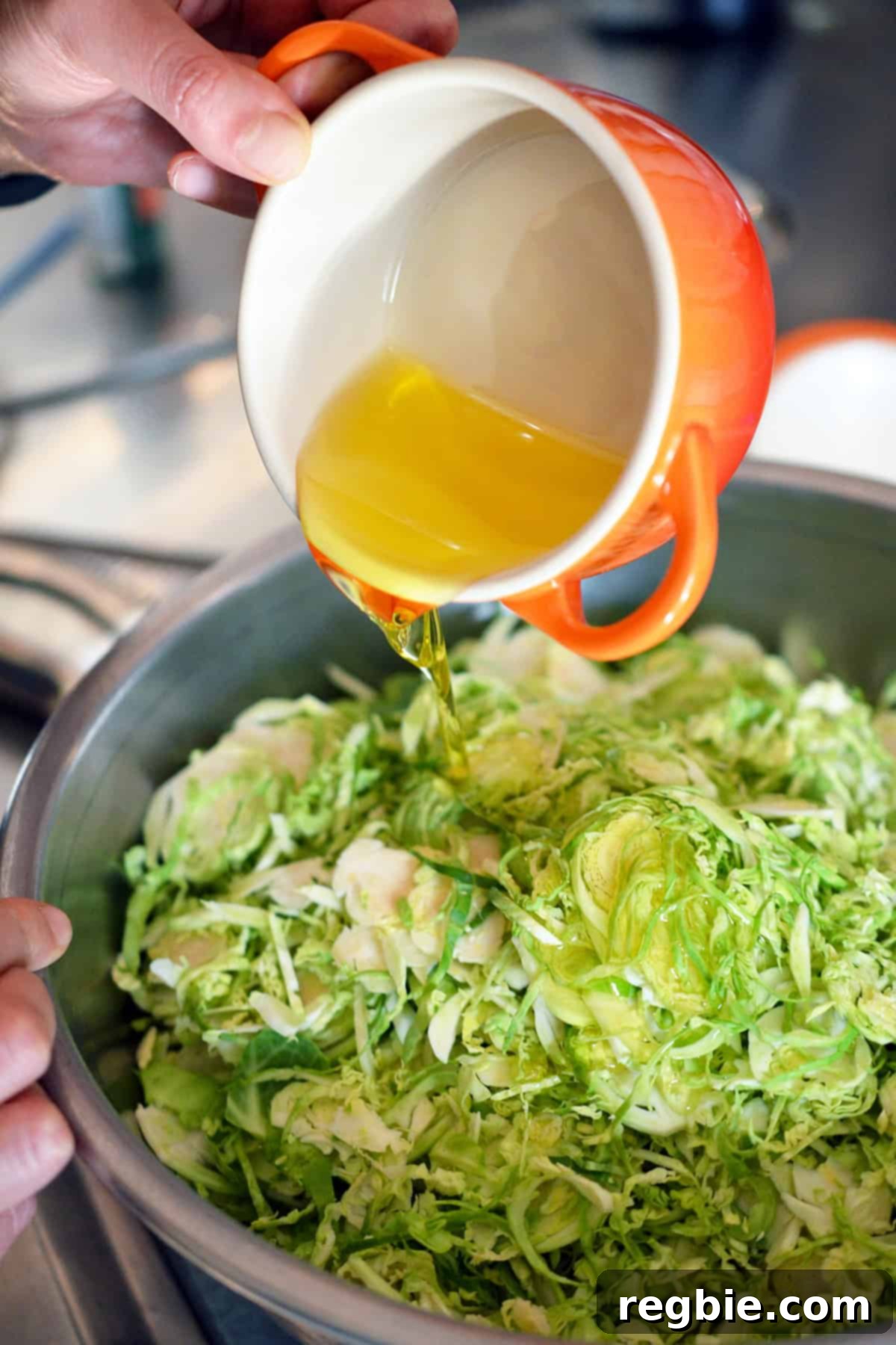 A small orange ramekin pouring melted ghee into a bowl of shredded raw Brussels sprouts, ready for seasoning and roasting.