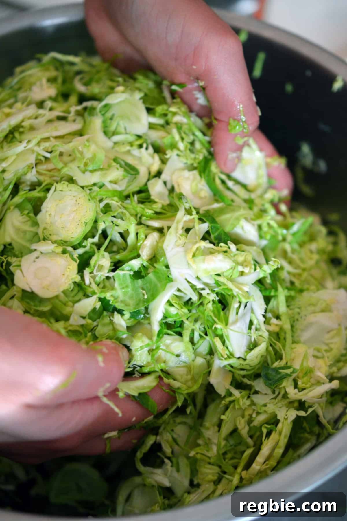 Hands actively tossing shaved Brussels sprouts with melted ghee and seasonings in a large metal bowl, ensuring every piece is coated for optimal roasting.
