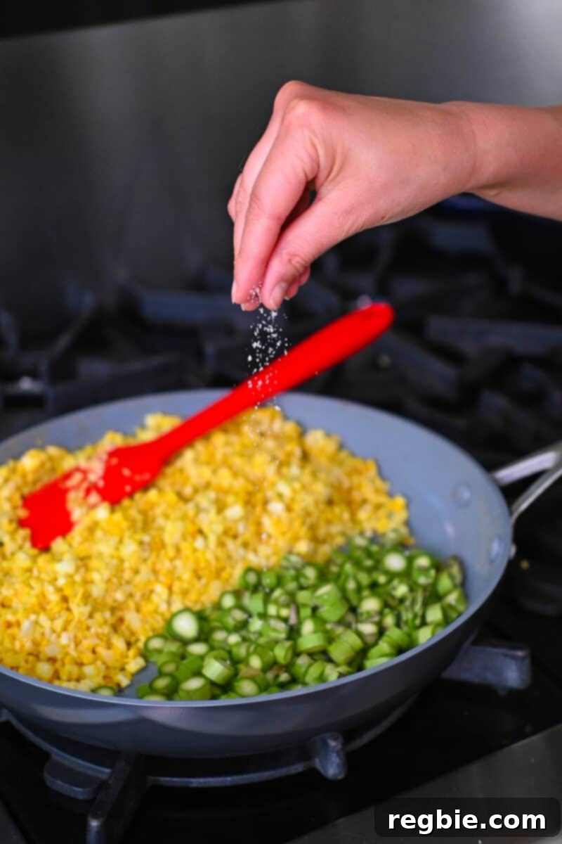 A hand is sprinkling salt on asparagus coins in a skillet, positioned next to the golden cauliflower fried rice, ready for cooking.