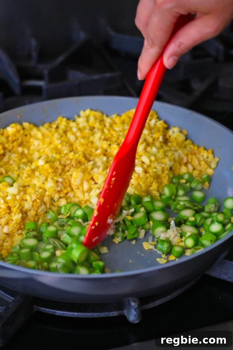 A red silicone spatula is mixing the vibrant asparagus coins with minced garlic in a pan that has yellow cauliflower rice in half of it, combining flavors.