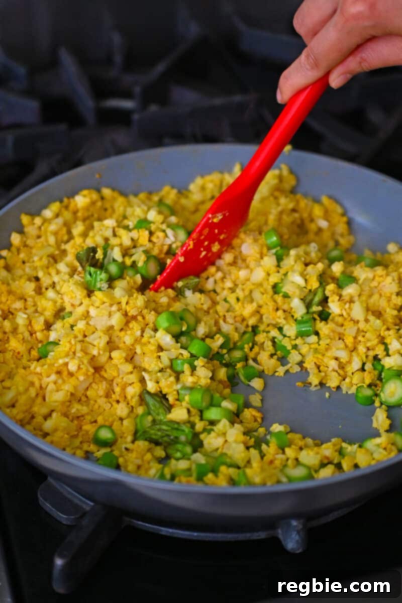 A red silicone spatula is stirring vibrant asparagus coins into the golden cauliflower fried rice, ensuring an even distribution of ingredients.