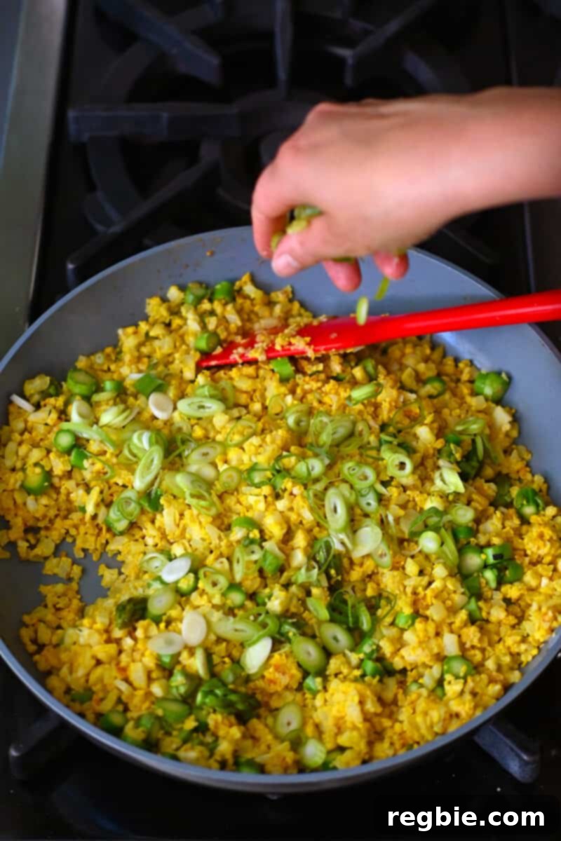A hand is sprinkling freshly sliced green onions onto a skillet filled with golden cauliflower fried rice, adding a final touch of freshness.