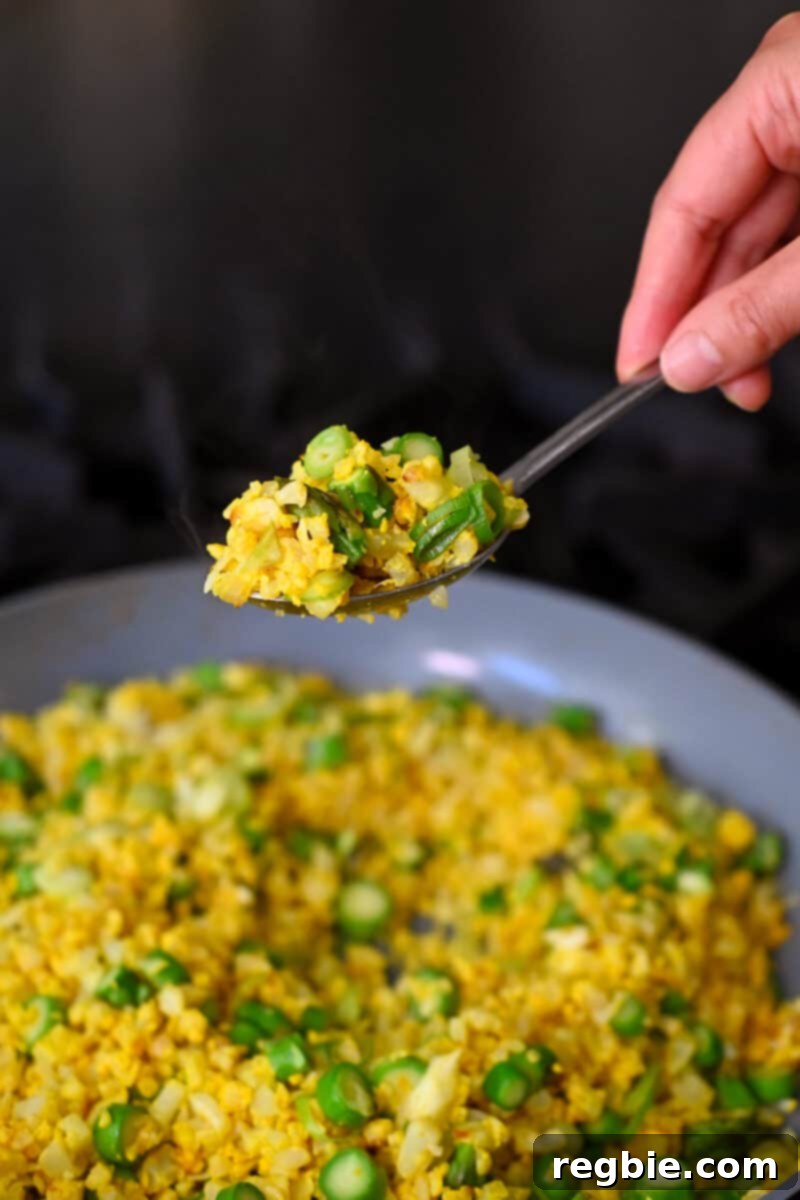 A spoon lifting up a scoop of golden cauliflower fried rice from a skillet, showcasing its vibrant color and fluffy texture.