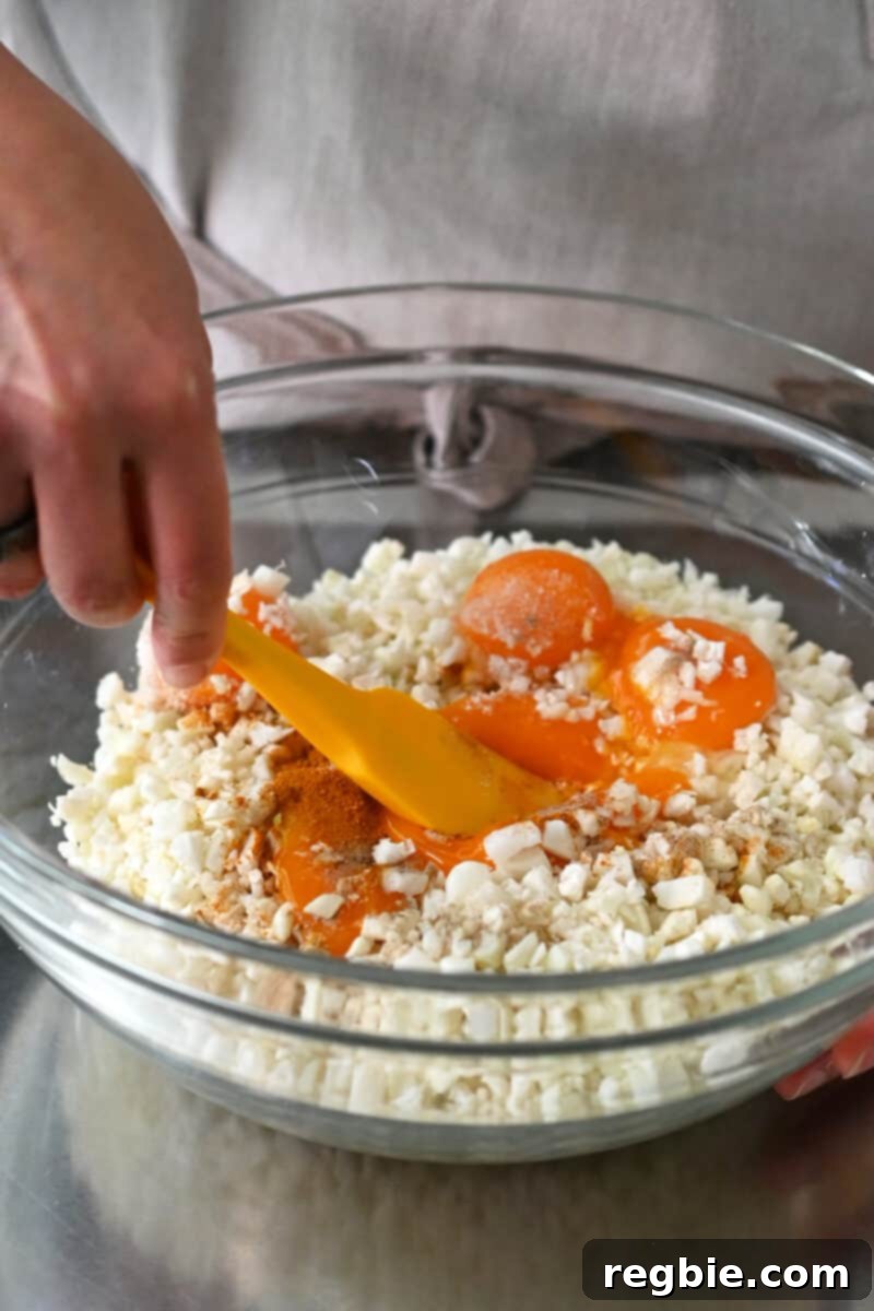 Combining finely chopped cauliflower rice in a large bowl with vibrant egg yolks and savory seasoning to create golden cauliflower fried rice.