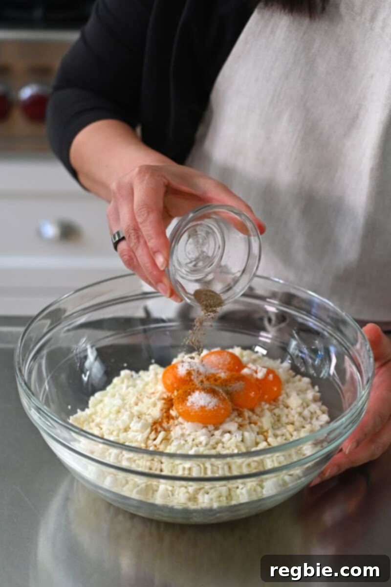 Ground white pepper is added to a large bowl filled with cauliflower rice, egg yolks, and salt, ready for mixing.