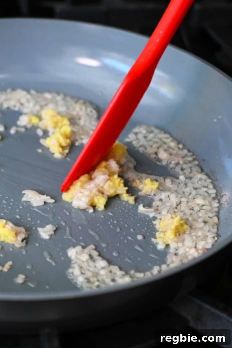 A closeup of a red silicone spatula pushing minced ginger and finely diced shallots in a large skillet, releasing their enticing aroma.