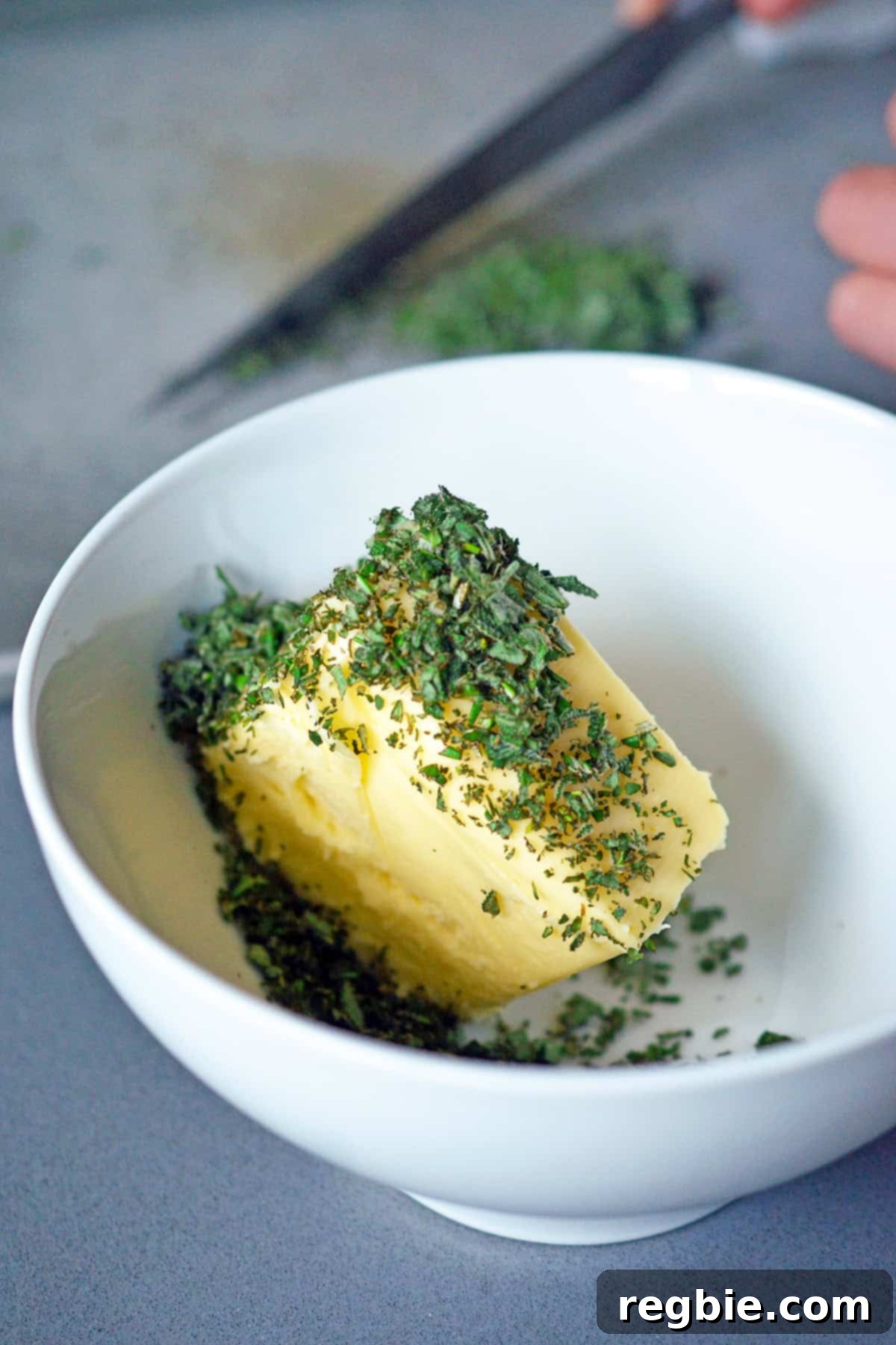 A white bowl containing softened butter topped with freshly minced herbs, ready for mixing.