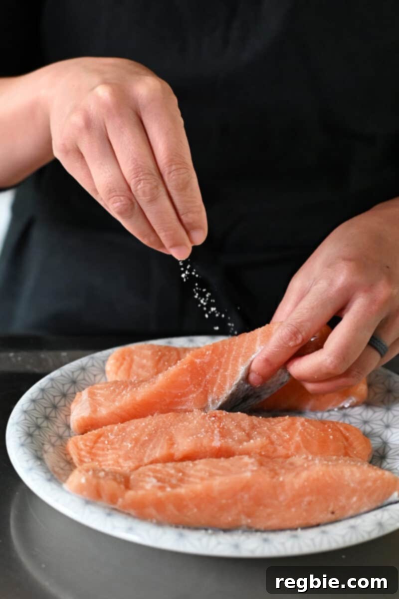 A hand sprinkles kosher salt onto four raw salmon fillets arranged neatly on a white plate, ready for seasoning.