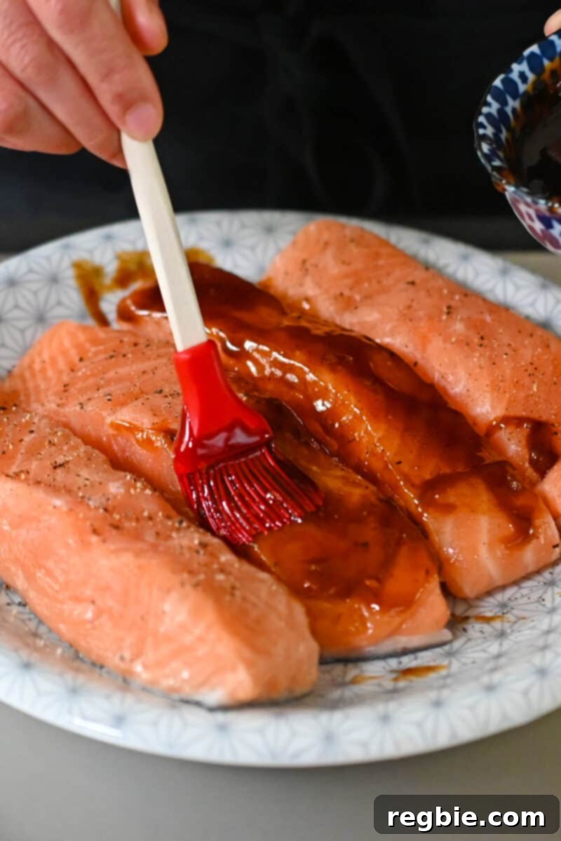 A silicone brush applies the rich honey sriracha sauce onto raw salmon fillets, ensuring they are evenly coated.