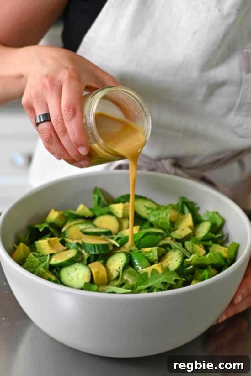 Whole30 Vegan Asian Chopped Salad 3 A hand pouring rich, amber-colored sesame ginger dressing into a large white bowl filled with a colorful medley of chopped salad ingredients.