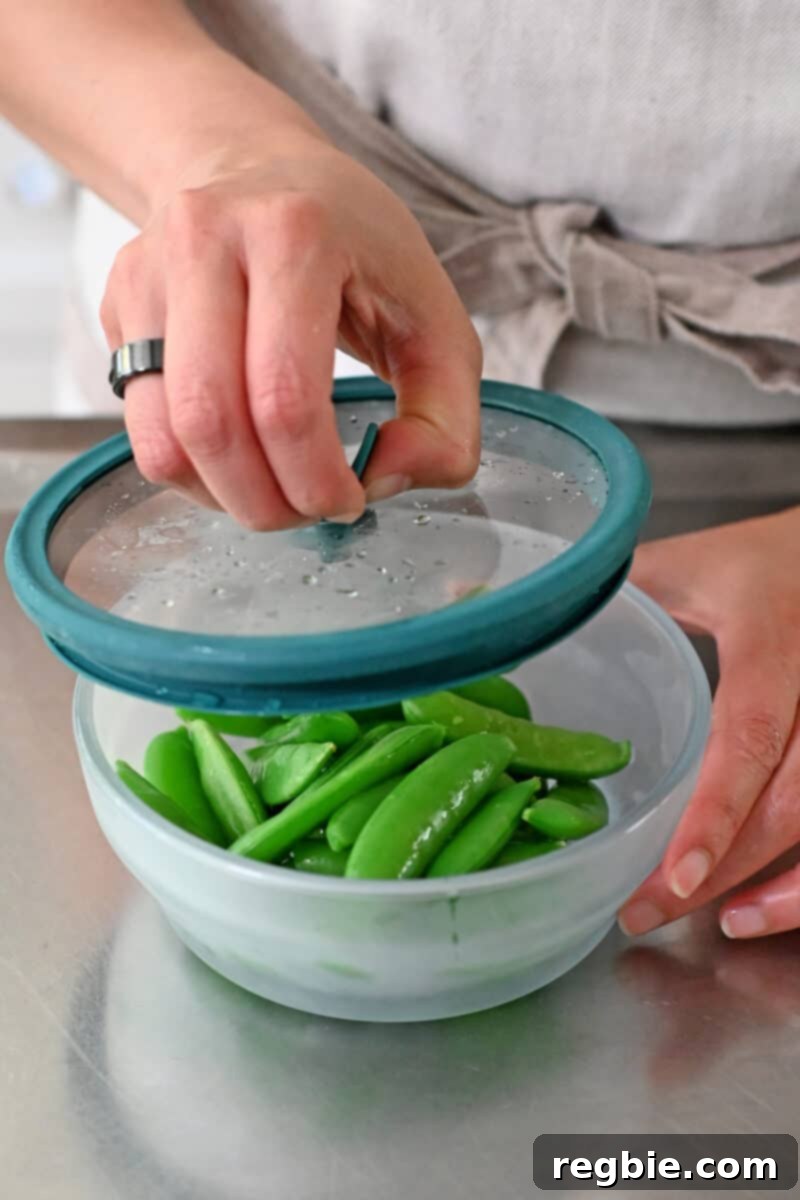 Whole30 Vegan Asian Chopped Salad 8 A hand opening a glass microwave container, revealing perfectly cooked, bright green sugar snap peas inside, ready for the next step.